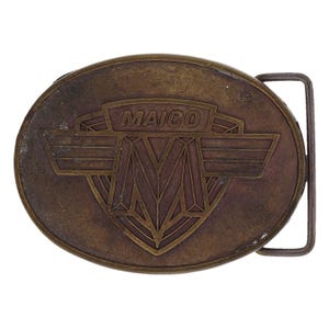 Puede incluir: Hebilla de cinturón ovalada de latón antiguo con la palabra "MAICO" en una pancarta rectangular. La hebilla presenta una "M" estilizada dentro de un diseño de escudo, flanqueada por alas. La hebilla tiene una barra rectangular para el cinturón.