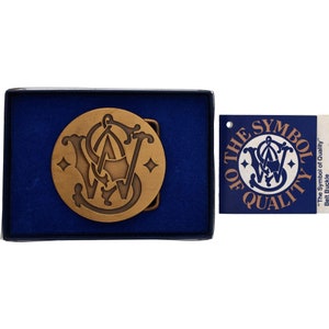 Op de afbeelding: Een gouden gesp met het Smith & Wesson-logo in een cirkelvormig ontwerp. De gesp zit in een blauwe fluwelen doos. De doos bevat een witte kaart met de tekst "The Symbol of Quality" en "Belt Buckle".