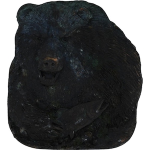 May include: A dark, textured sculpture of a bear holding a fish. The bear is black with bronze-coloured accents on its snout and mouth. The fish is also black. The bear's expression is fierce, with its mouth open.