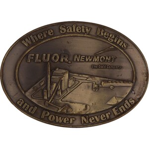 Può includere: Una fibbia per cintura ovale in bronzo con il testo "Where Safety Begins and Power Never Ends" attorno al bordo. Il centro della fibbia presenta una rappresentazione di una fabbrica con il testo "Fluor. Newmont The Gold Company".