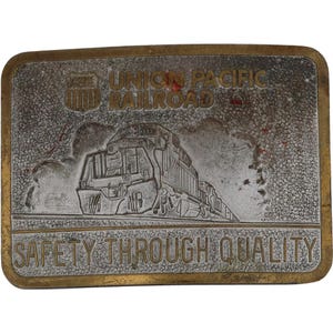 Op de afbeelding: Rechthoekige metalen riemgesp met een goudkleurige rand. De gesp toont de woorden "UNION PACIFIC RAILROAD" en "SAFETY THROUGH QUALITY" in gouden letters. Een gedetailleerde afbeelding van een trein staat in het midden.