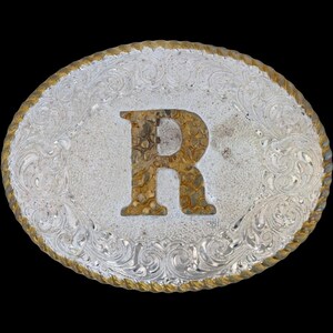 May include: Oval silver-tone belt buckle with a gold-colored rope edge. The center features a large letter "R" in a textured, brown and gold pattern. The silver portion has an ornate, swirling design.