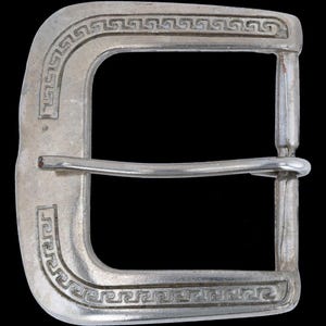 Puede incluir: Hebilla de cinturón plateada con forma rectangular y un patrón decorativo de grecas en los bordes. La hebilla tiene una barra para la correa y un pasador para el cierre.