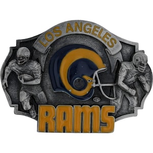 May include: A silver-toned metal belt buckle featuring the Los Angeles Rams logo. The buckle has a football helmet in the center, with the words "LOS ANGELES" and "RAMS" in gold lettering. Two football players are on either side.