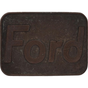 May include: A rectangular, bronze-colored metal plaque with rounded corners. The word "Ford" is embossed in a matching bronze color across the center of the plaque. The surface has a weathered appearance.