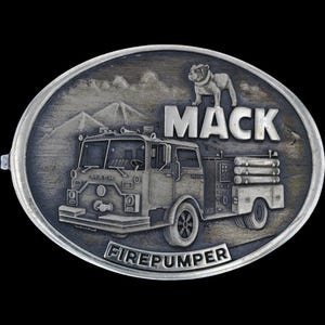 May include: Oval silver-coloured belt buckle with a detailed relief of a Mack fire truck, a bulldog, and the word "MACK." The word "FIREPUMPER" is inscribed at the bottom. Mountains and clouds are in the background.