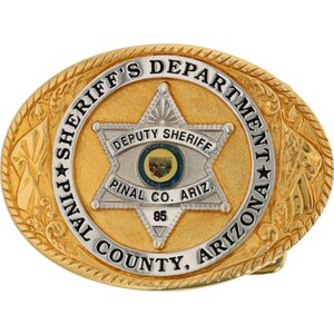 May include: An oval gold-tone belt buckle with the words "SHERIFF'S DEPARTMENT" and "PINAL COUNTY, ARIZONA" in raised lettering. The centre features a silver star with "DEPUTY SHERIFF" and "PINAL CO. ARIZ." The number "95" is at the bottom.