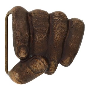May include: A bronze-coloured belt buckle in the shape of a clenched fist. The buckle has a rectangular loop on the left side for the belt. The fist is detailed with knuckles and a textured surface.