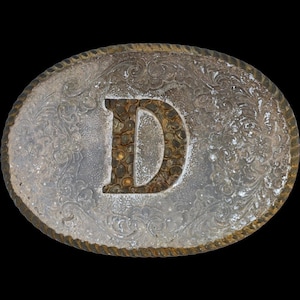 Può includere: Fibbia per cintura ovale color argento con un motivo floreale decorativo e un bordo a forma di corda. La lettera "D" è al centro, con un riempimento strutturato color marrone. La fibbia è su sfondo nero.