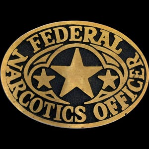 Puede incluir: Insignia ovalada de metal dorado con las palabras "NARCOTICS OFFICER FEDERAL" en relieve. El centro presenta una estrella grande y dos estrellas más pequeñas, todas doradas, sobre un fondo negro.