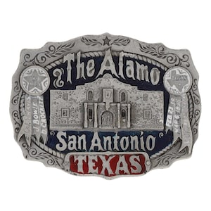 May include: A silver-toned metal belt buckle featuring a detailed depiction of The Alamo in San Antonio, Texas. The buckle includes text that reads "The Alamo", "San Antonio", and "Texas". Ribbons and stars are also included in the design.