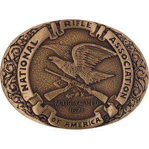 Puede incluir: Hebilla de cinturón ovalada de bronce con un águila con las alas extendidas, rifles cruzados y el texto "NATIONAL RIFLE ASSOCIATION INCORPORATED 1871 OF AMERICA". La hebilla tiene un borde decorativo.