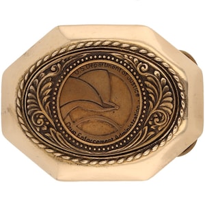 Pode incluir: Uma fivela de cinto de metal dourado com formato octogonal. A fivela apresenta um design gravado de uma águia estilizada com o texto "U.S. Department of Justice Drug Enforcement Administration".