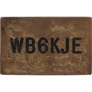 Puede incluir: Placa rectangular de latón envejecido con el texto en relieve "WB6KJE" en negro. La placa tiene un aspecto moteado y envejecido, con variaciones de color y textura en su superficie.