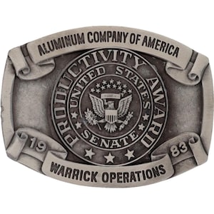 Pode incluir: Uma fivela de cinto de metal prateado com o texto "ALUMINUM COMPANY OF AMERICA" e "PRODUCTIVITY UNITED STATES SENATE AWARD". A fivela também apresenta o texto "WARRICK OPERATIONS" e os números "19" e "83".