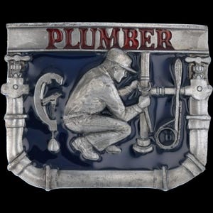 Może przedstawiać: Prostokątna metalowa klamra do paska z niebieskim tłem i napisem "PLUMBER" w czerwonych literach. Klamra przedstawia srebrny relief hydraulika pracującego przy rurach. Klamra ma srebrną obwódkę.