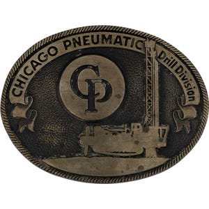 May include: Bronze-toned, oval belt buckle with the text "CHICAGO PNEUMATIC Drill Division." The centre circle features the letters "CP" and a drilling rig graphic. The buckle has a textured surface and a decorative border.