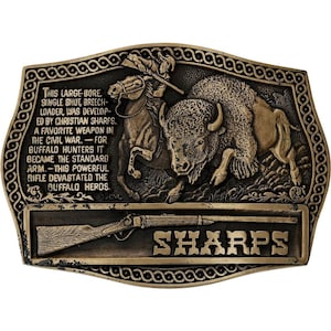 May include: A rectangular, bronze-colored belt buckle with a detailed relief scene. It depicts a mounted figure shooting at a buffalo, with text that reads "SHARPS" and historical information about the rifle.
