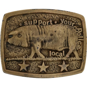 Peut inclure: Boucle de ceinture rectangulaire en laiton antique avec une illustration de cochon. La boucle porte l'inscription "Support Your Local Police" et comporte trois étoiles en bas. Le cochon marche sur l'herbe.