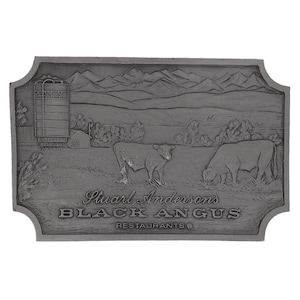 Op de afbeelding: Een rechthoekige, grijze metalen plaat met een gedetailleerde reliëfscène. Het toont twee koeien in een veld, een silo, een klein gebouw en bergen op de achtergrond. De tekst "Stuart Andersons BLACK ANGUS RESTAURANTS®" is gegraveerd.