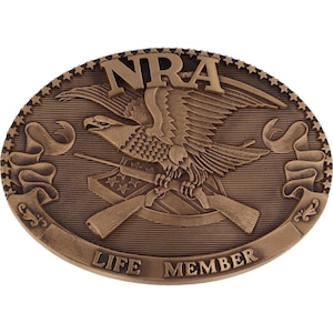 Puede incluir: Una hebilla de cinturón de bronce con un águila sosteniendo rifles cruzados y una bandera estadounidense. La hebilla tiene el texto "NRA LIFE MEMBER" alrededor del borde.