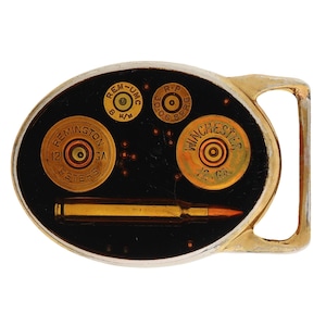 May include: Oval-shaped belt buckle with a gold-colored frame and a black background. The buckle features several brass-colored bullet casings and a bullet. The casings are labeled "Remington Peters" and "Winchester".