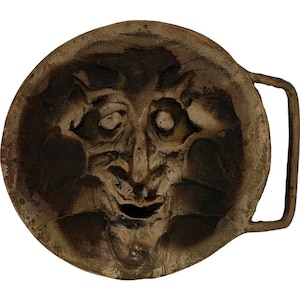 May include: A vintage, circular belt buckle with a sculpted face design. The buckle is made of a dark, weathered metal with a detailed, expressive face in the center. The buckle has a rectangular bar for the belt.