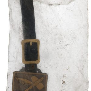 May include: A vintage brown and gold metal tag with a black strap and buckle, featuring the word "DROTT" and a crossed axe and wing design. The tag is enclosed in a translucent paper envelope.