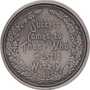 May include: A silver-colored coin with the inscription "Success Comes to Those Who Hustle Wisely." The text is in a stylized font, surrounded by a decorative wreath. The coin has a detailed border.