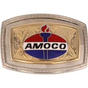 Puede incluir: Una hebilla de cinturón de plata y oro con un logotipo de forma ovalada que presenta la palabra "AMOCO" en letras blancas sobre un fondo azul y rojo. Una llama estilizada se encuentra sobre el logotipo.