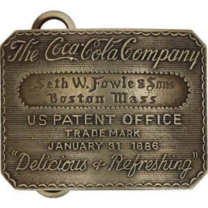 May include: A rectangular, antique-style metal plaque with the text "The Coca-Cola Company" at the top. The plaque also features the text "Seth W. Fowle & Sons Boston Mass", "US PATENT OFFICE", "TRADE MARK", "JANUARY 31 1886", and "Delicious & Refreshing".