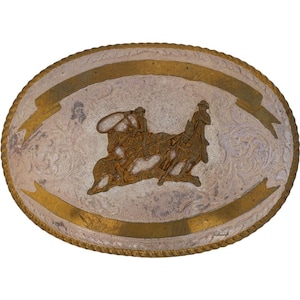 Puede incluir: Hebilla de cinturón ovalada plateada con borde y detalles dorados. La hebilla presenta un relieve detallado de dos vaqueros a caballo lazando un toro. El fondo tiene un patrón floral grabado.
