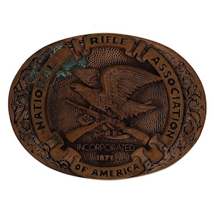 May include: Oval-shaped, bronze-colored belt buckle featuring an eagle with crossed rifles. The buckle is embossed with the text "National Rifle Association of America, Incorporated 1871". Ornate scrollwork frames the central design.