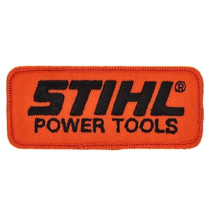 Könnte beinhalten: Ein orangefarbener rechteckiger Aufnäher mit dem schwarzen Text "STIHL POWER TOOLS". Der Aufnäher hat einen genähten Rand und ist wahrscheinlich ein Emblem für Arbeitskleidung oder Geräte.