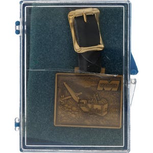 Könnte beinhalten: Eine Vintage-Gürtelschnalle mit einem goldfarbenen rechteckigen Rahmen und einem schwarzen Lederriemen. Die Schnalle wird in einer transparenten Kunststoffbox mit türkisem Hintergrund präsentiert. Die Schnalle trägt das Wort "MARION" und ein Bild eines Krans.