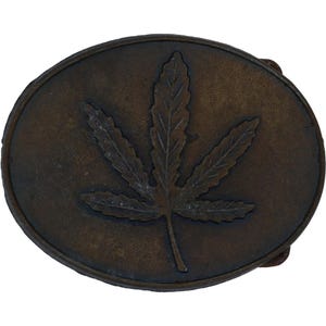 May include: Oval-shaped, bronze-colored belt buckle featuring a detailed cannabis leaf design. The leaf is centrally positioned, with intricate veining, set against a textured background. The buckle has a classic, rustic appearance.