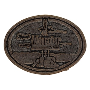 Puede incluir: Hebilla de cinturón ovalada de color bronce con la palabra "Magcobar" en el centro. La hebilla presenta un relieve detallado de una plataforma petrolífera y un paisaje. La hebilla es de metal y tiene una superficie texturizada.