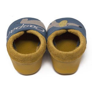May include: A pair of blue baby shoes with a yellow lining and a gray and gold embroidered animal design. The left shoe has the word "Cospa" embroidered on it.