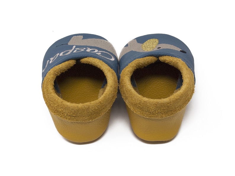 May include: A pair of blue baby shoes with a yellow lining and a gray and gold embroidered animal design. The left shoe has the word "Cospa" embroidered on it.