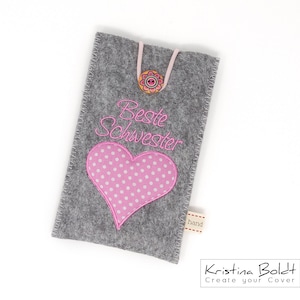 Könnte beinhalten: Grauer Filz-Handy-Etui mit einem rosa Herz und dem gestickten Text "Beste Schwester". Das Etui hat einen rosa Knopf und ein kleines Etikett mit der Aufschrift "hand".