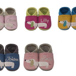 May include: Ten pairs of baby shoes with a dachshund design. The shoes are in various colors, including pink, green, blue, and yellow. Each pair has a different name embroidered on them, including Clara, Lilly, Lea, Johann, and Lima.