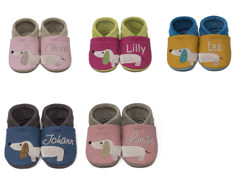 May include: Ten pairs of baby shoes with a dachshund design. The shoes are in various colors, including pink, green, blue, and yellow. Each pair has a different name embroidered on them, including Clara, Lilly, Lea, Johann, and Lima.