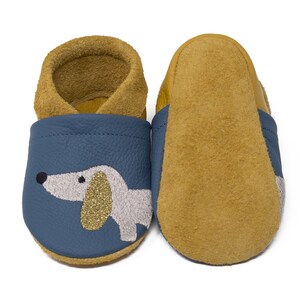 May include: A pair of blue leather baby shoes with a gold glitter dachshund design. The shoes have a brown suede lining and a brown suede sole.