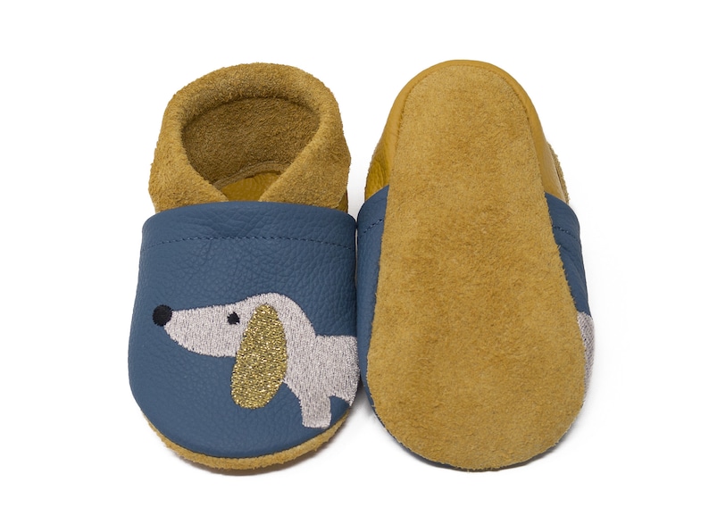 May include: A pair of blue leather baby shoes with a gold glitter dachshund design. The shoes have a brown suede lining and a brown suede sole.