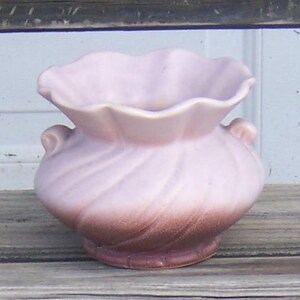 May include: A small, vintage, pink ceramic vase with a ruffled rim and two small handles. The vase has a two-tone color scheme, with a lighter pink at the top that fades into a darker brown at the base. The vase has a textured, swirled design.