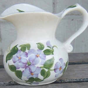 May include: A white ceramic pitcher featuring a hand-painted floral design with light purple petals, yellow centers, and green leaves. The pitcher has a rounded body, a curved handle, and a wide, scalloped pouring spout at the top rim.