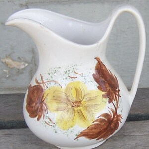 May include: A white ceramic pitcher featuring a hand-painted floral design. The central motif is a large yellow flower with brown brushstroke petals and leaves, accented by small green speckled details. The pitcher has a curved handle and a spout.