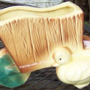 Könnte beinhalten: Ein Keramik-Pflanzgefäß in Form einer Schubkarre mit Holzmaserung in Beige- und Brauntönen. Das Rad ist grün bemalt und eine kleine, hellgelbe Entenfigur ist an der Vorderseite der Schubkarrenstruktur angebracht.