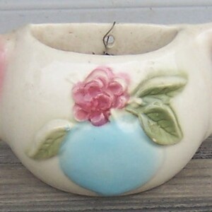 Könnte beinhalten: Vintage Keramik-Blumentopf in Teekannenform, cremefarben mit rosa Akzenten an Ausguss und Henkel. Er hat ein erhabenes Blumendesign mit einer rosa Rose und einem herzförmigen Detail in Blau. Ein dekorativer Artikel.