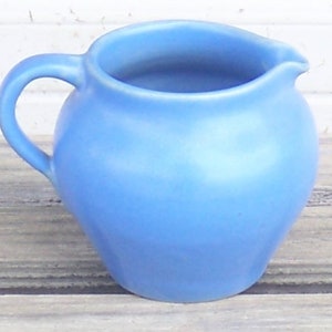 May include: A small, round, light blue ceramic creamer with a handle and a pouring spout. The creamer has a smooth, matte finish and sits on a wooden surface. The handle is curved and attached to the side of the creamer.